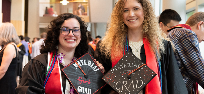Two students in graduation gowns, holding their decorated caps that say Materials Grad 